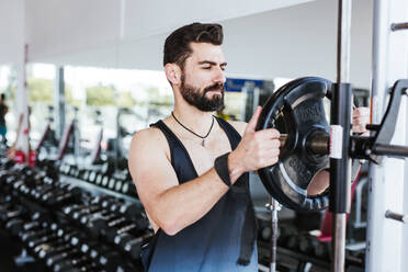 Muscular bearded man in sportswear standing near squat rack and putting weight disk on barbell while preparing for weightlifting training in modern gym - ADSF13501