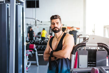 Muskulöser, bärtiger Mann in Sportkleidung schaut in die Kamera, während er mit verschränkten Armen auf einem Sportgerät in einem hellen, modernen Fitnessstudio steht - ADSF13502