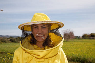 Positive Imkerin in gelbem Schutzanzug und Maske, die lächelnd in die Kamera schaut, während sie auf einer grünen Wiese in einem Bienenstock an einem sonnigen Sommertag steht - ADSF13510