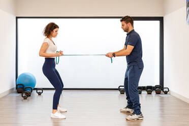 Seitenansicht von männlichen und weiblichen Sportlern in aktiver Kleidung, die voreinander stehen und an einem Gummiband in einem modernen Fitnessstudio ziehen - ADSF13524