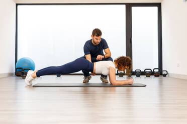 Side view of strong woman in plank position with male personal trainer in modern gym - ADSF13525