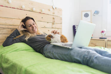 Lächelnde Frau, die ein Meerschweinchen streichelt, während sie mit ihrem Laptop zu Hause auf dem Bett liegt - SKCF00680