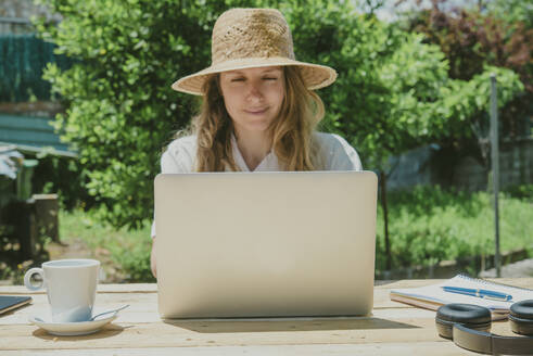 Lächelnde Frau bei der Arbeit, während sie am Tisch im Hinterhof sitzt und einen Laptop benutzt - SKCF00692
