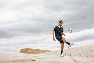 Junger sportlicher Mann in schwarzem T-Shirt und Shorts, der beim Training auf einer gepflasterten Straße mit bewölktem Himmel im Hintergrund einen Fußball kickt - ADSF13541