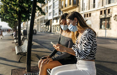 Young friends with face mask using phone while sitting on bench at promenade - MGOF04378