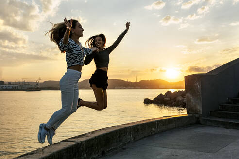 Junge Frauen, die bei Sonnenuntergang auf der Promenade springen - MGOF04395