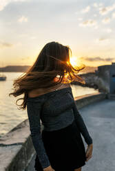 Frau, die sich die Haare zuwirft, während sie bei Sonnenuntergang an der Promenade steht - MGOF04398