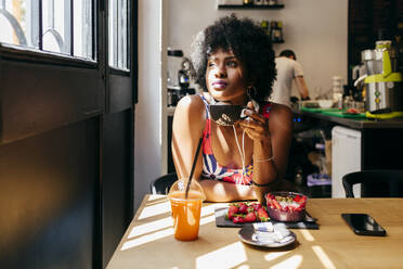 Schöne junge Afroamerikanerin trinkt Kaffee und schaut nachdenklich zum Fenster, während sie ein gesundes Frühstück mit Erdbeer-Smoothie und Fruchtsaft in einem kleinen Café zu sich nimmt - ADSF13591