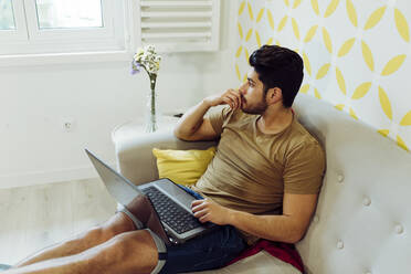 Side view of young thoughtful man in casual outfit with laptop sitting on couch near table with fresh flowers - ADSF13593