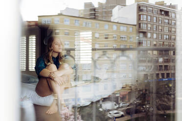 Junge Frau sitzt auf dem Bett und schaut durch das Fenster - ADSF13599