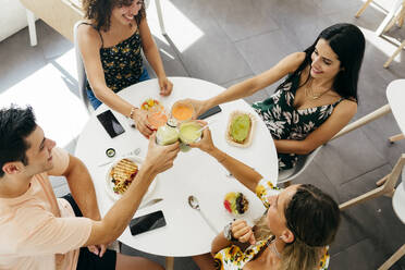 Multiethnische junge Leute stoßen mit Gläsern frischer Fruchtgetränke an und bringen Toasts aus, während sie in einem Café mit gesunden Snacks am Tisch sitzen - ADSF13601