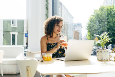 Ethnische Frau mit lockigem Haar, die einen frischen Saft trinkt und einen Laptop benutzt, während sie in einem modernen, stilvollen Restaurant am Fenster sitzt - ADSF13607