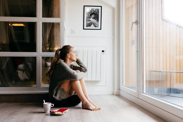 Side view of calm young female in casual wear looking at window thoughtfully while sitting on floor with cup of coffee and toasts during breakfast at home - ADSF13625