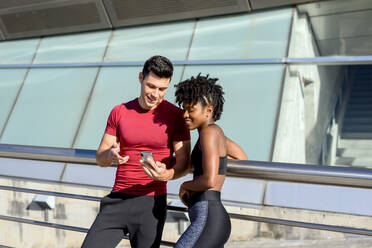 Personal trainer using smartphone and showing app to smiling fitness African American female while standing together next to building in sunny summer day - ADSF13626