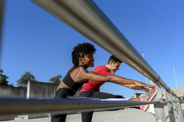 Von unten Seitenansicht der fröhlichen afroamerikanischen Fitness-Frau und des lächelnden sportlichen Mannes, die neben einem Metallzaun stehen und sich die Beine vertreten, während sie gemeinsam an einem sonnigen Tag mit blauem Himmel im Hintergrund trainieren - ADSF13627
