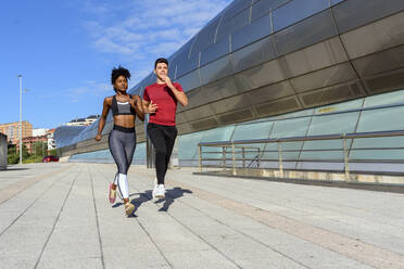 Junge sportliche afroamerikanische Frau in Top und Leggings joggt mit einem sportlichen männlichen Freund neben einer modernen Stadtanlage im Sommer - ADSF13628