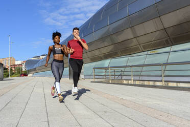 Young athletic African American female in top and leggings jogging with sportive male friend next to modern city construction in summer day - ADSF13628