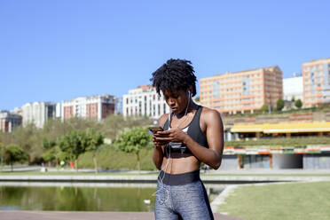 Junge aktive Afroamerikanerin in Sportkleidung hört mit Smartphone und Kopfhörern Musik beim Training am Flussufer in der Stadt - ADSF13630