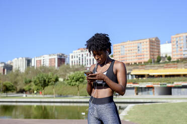 Young active African American female in sportswear listening to music with smartphone and earphones while training at river bank in city - ADSF13630