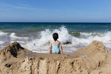 Back view of unrecognizable little girl in swimsuit sitting on sandy beach against sea waves and enjoying summer holidays in sunny day - ADSF13631