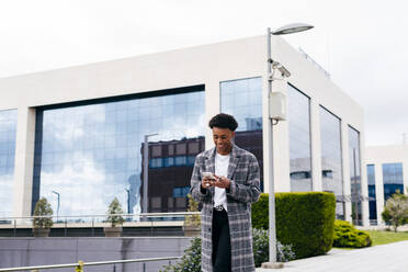 From below of happy young African American male student in stylish outfit browsing smartphone while standing on city street near modern building - ADSF13634