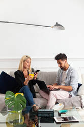 Positive blonde woman with cup of juice browsing smartphone and sitting on couch near ethnic boyfriend typing on laptop keyboard in living room of modern apartment - ADSF13649