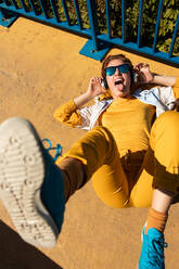 Von oben trendy Chillen Teenager-Frau in Sonnenbrille und leuchtend gelbe Kleidung Musik hören mit drahtlosen Kopfhörern, während auf lebendige Bürgersteig mit blauen Zaun neben in sonnigen Tag liegen - ADSF13655