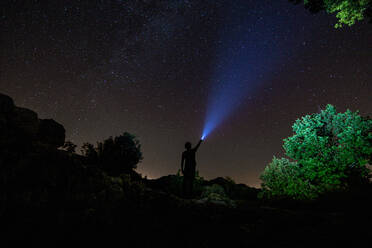 Silhouette einer Person mit einer Taschenlampe, die den Himmel und die Sterne im Dunkeln auf einem grünen Platz beleuchtet - ADSF13667