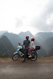 Seitenansicht eines glücklichen Paares von Reisenden, die auf einem am Straßenrand geparkten Motorrad sitzen und sich vor dem Hintergrund einer Bergkette ansehen - ADSF13696
