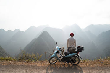 Back view of traveling man wearing protective helmet standing near motorbike and admiring picturesque scenery of mountain range in misty morning - ADSF13697