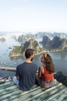 Von oben Rückansicht des liebenden Paares, das auf dem Metalldach sitzt und die malerische Aussicht auf die Halong-Bucht in Vietnam betrachtet - ADSF13700