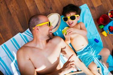 From above view of cheerful boy in yellow sunglasses and blue swimwear smiling while feeding father apple lying on towels together having beach at home - ADSF13714