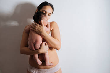 Happy pregnant woman kneeling on the floor in a studio, smiling at her  beautiful baby bump. Young mom-to-be wearing lingerie, cherishing the life  growing inside her with maternal love. stock photo