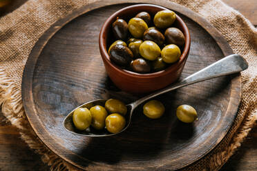 From above wooden plate bowl and spoon with black and green olives placed on rustic fabric on wooden table - ADSF13739