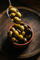 Draufsicht auf einen Metalllöffel mit Oliven in Öl vor dem Hintergrund einer Keramikschale auf einem Holztisch in der Küche - ADSF13741