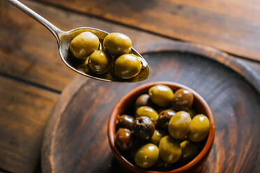 Draufsicht auf einen Metalllöffel mit Oliven in Öl vor dem Hintergrund einer Keramikschale auf einem Holztisch in der Küche - ADSF13742