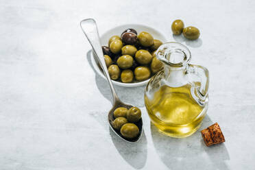 Von oben: Glaskanne mit Öl und kleine Schale und Löffel mit Oliven auf weißem Tisch - ADSF13743