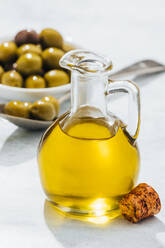 Glaskanne mit Öl und kleine Schale und Löffel mit Oliven auf weißem Tisch - ADSF13744