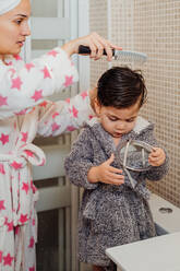 Side view of mother wearing bathrobe and towel turban combing wet hair of little kid while standing in modern bathroom - ADSF13759