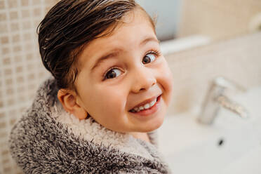 Von oben auf ein fröhliches kleines Kind mit nassen Haaren und weichem Bademantel, das neben dem Waschbecken im Badezimmer steht und in die Kamera schaut - ADSF13761