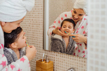 Cute little boy in bathrobe and smiling mother in towel turban standing in bathroom and brushing teeth - ADSF13763