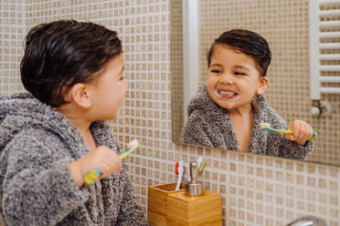 Adorable Kind trägt kuscheligen Bademantel stehend im Bad mit Zahnbürste und Blick in den Spiegel - ADSF13764