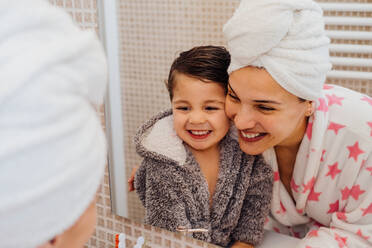 Fröhliche Frau mit Handtuch-Turban, die ein kleines Kind im Bademantel kuschelt, nachdem sie geduscht und in den Spiegel geschaut hat - ADSF13765