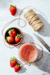 Composition with fresh homemade strawberry juice in glass jar wrapped with twine placed on marble surface with whole berries and knife - ADSF13775