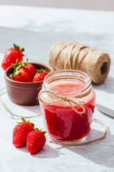 Komposition mit frischem, hausgemachtem Erdbeersaft in einem mit Bindfaden umwickelten Glasgefäß, das mit ganzen Beeren und einem Messer auf eine Marmorplatte gestellt wird - ADSF13776