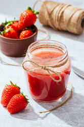 Komposition mit frischem, hausgemachtem Erdbeersaft in einem mit Bindfaden umwickelten Glasgefäß, das mit ganzen Beeren und einem Messer auf eine Marmorplatte gestellt wird - ADSF13777