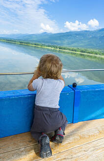 Kleiner Junge kniet vor einem Geländer und betrachtet die malerische Aussicht auf den Kerkini-See, Mazedonien, Griechenland - RUNF04116