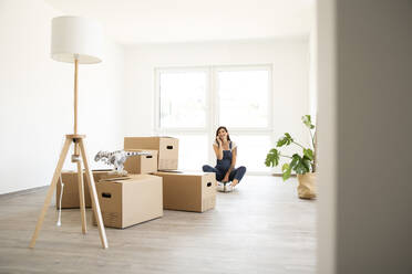 Pregnant woman talking over smart phone while sitting by cardboard boxes in new home - MJFKF00566