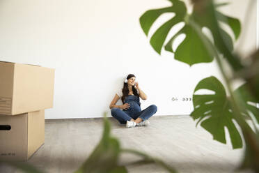 Pregnant woman listening music through headphones while sitting against wall in new home - MJFKF00573