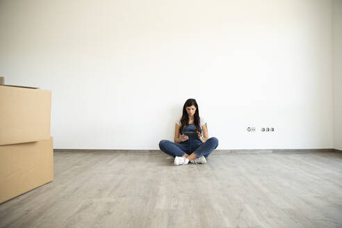 Pregnant woman using digital tablet while sitting on floor against white wall in new house - MJFKF00574